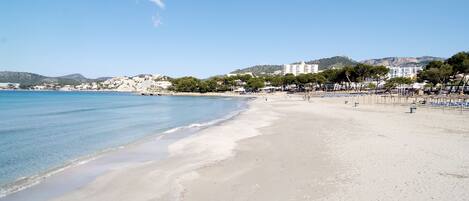 A beach with clear blue water, sandy shore, and buildings in the background.