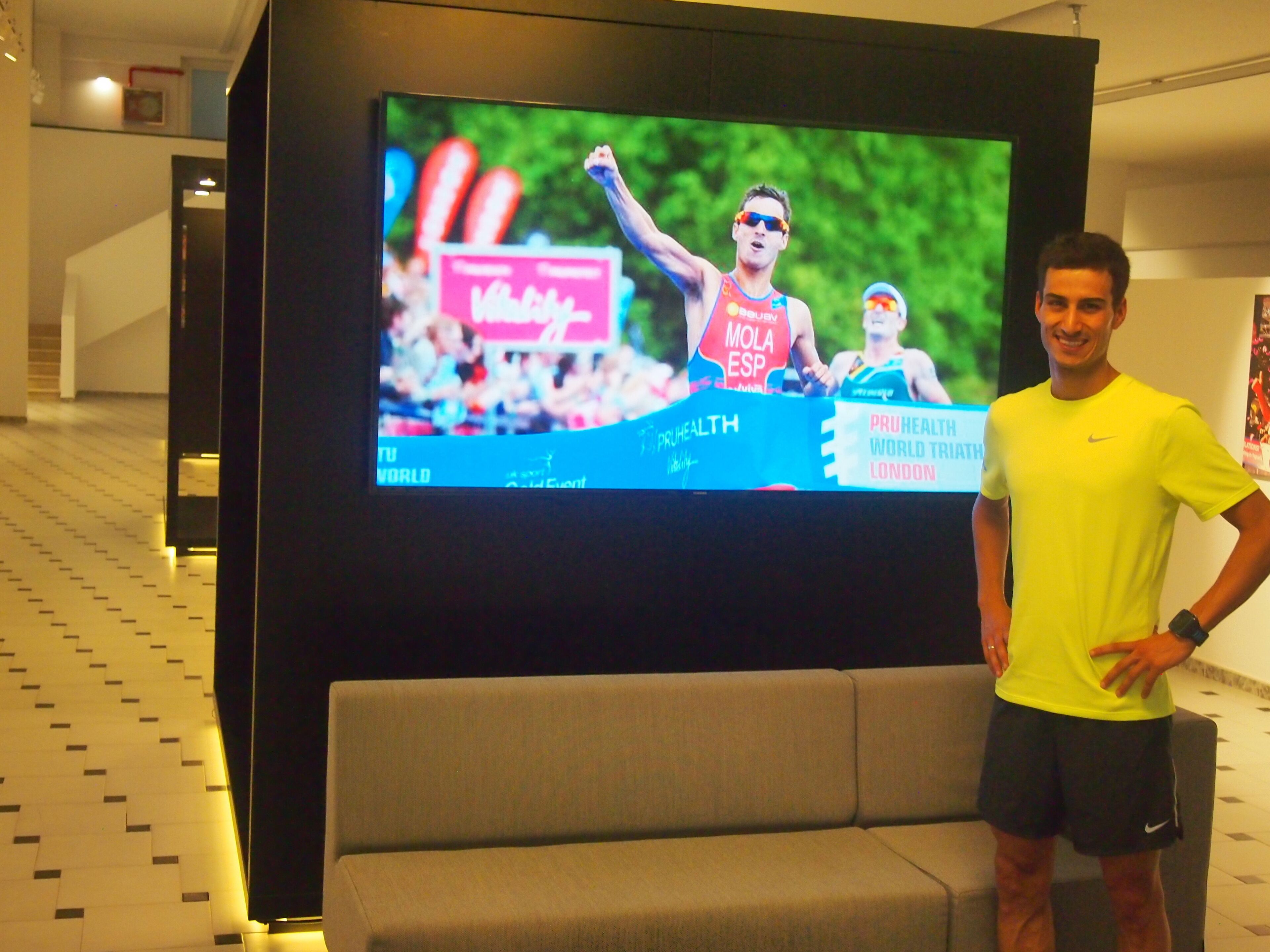 Un hombre de pie frente a un televisor que muestra un evento deportivo, con un sofá y un suelo estampado al fondo.