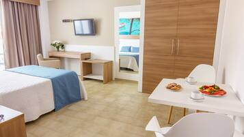 Habitación de hotel con cama, escritorio, silla, televisor y mesa de desayuno con croissants y fruta.