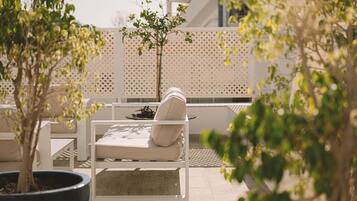 Un área de estar al aire libre iluminada por el sol, con una silla blanca acolchada y una mesa pequeña, rodeada de plantas en macetas y un cerramiento decorativo.