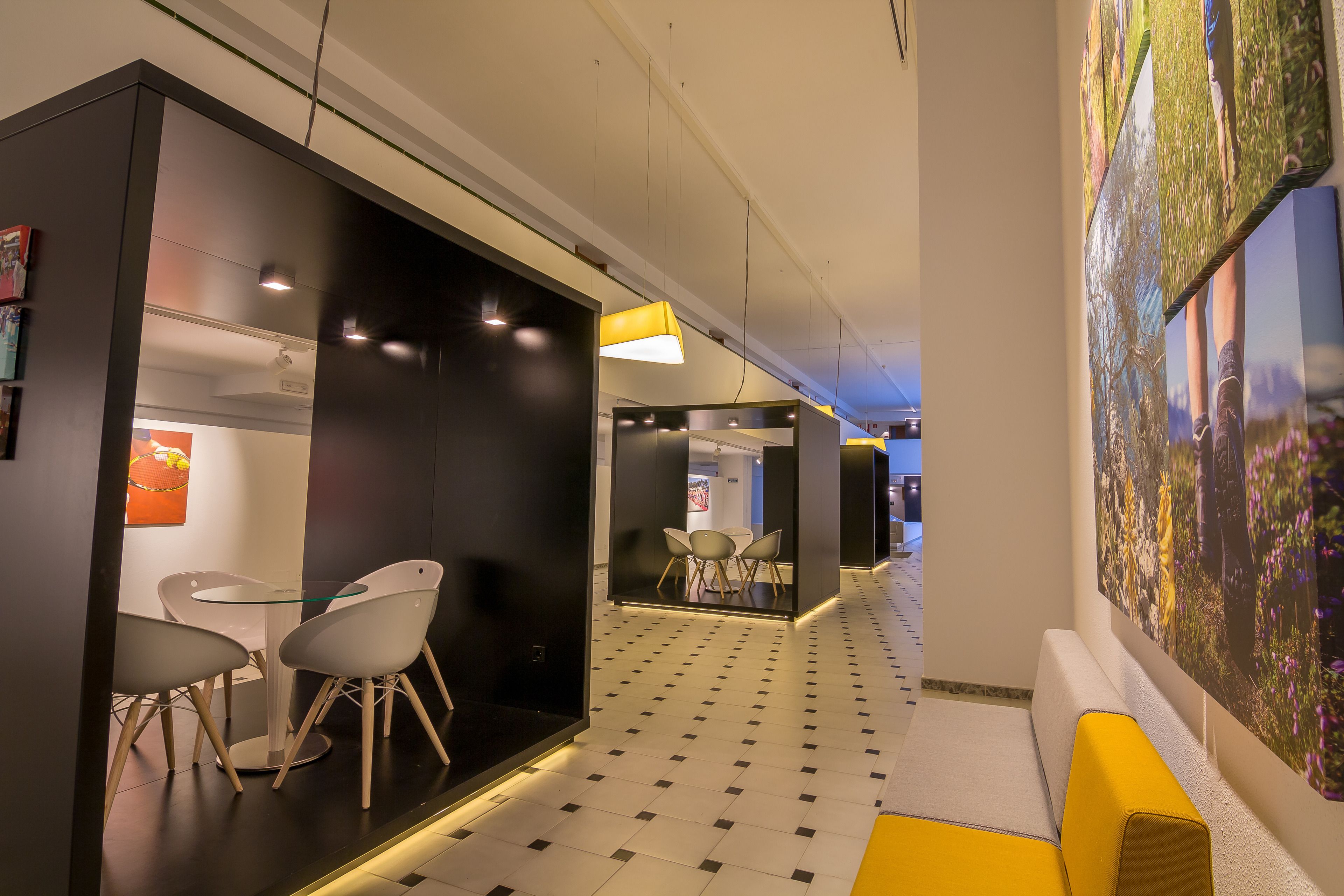 A modern hotel corridor with a checkered floor, a seating area with white chairs, and a large wall mural.