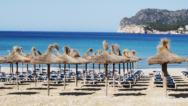 A beach with rows of blue lounge chairs and thatched umbrellas, a clear blue sea, and a rocky cliff in the background. - Aparthotel Novo Mar (Calvia)
