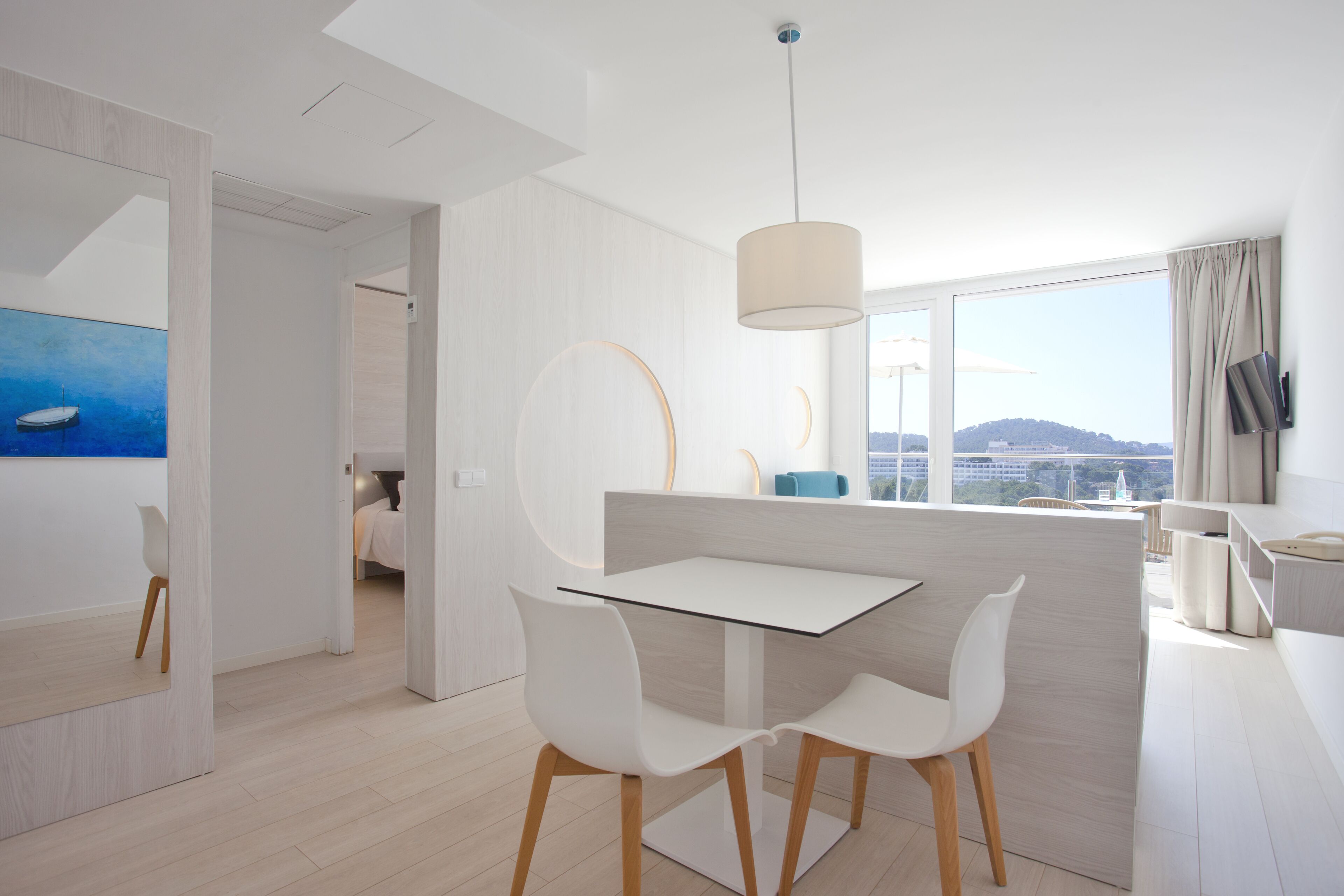 Una habitación de hotel moderna con una mesa de comedor de vidrio, sillas blancas, un ventanal con vistas y un diseño minimalista.