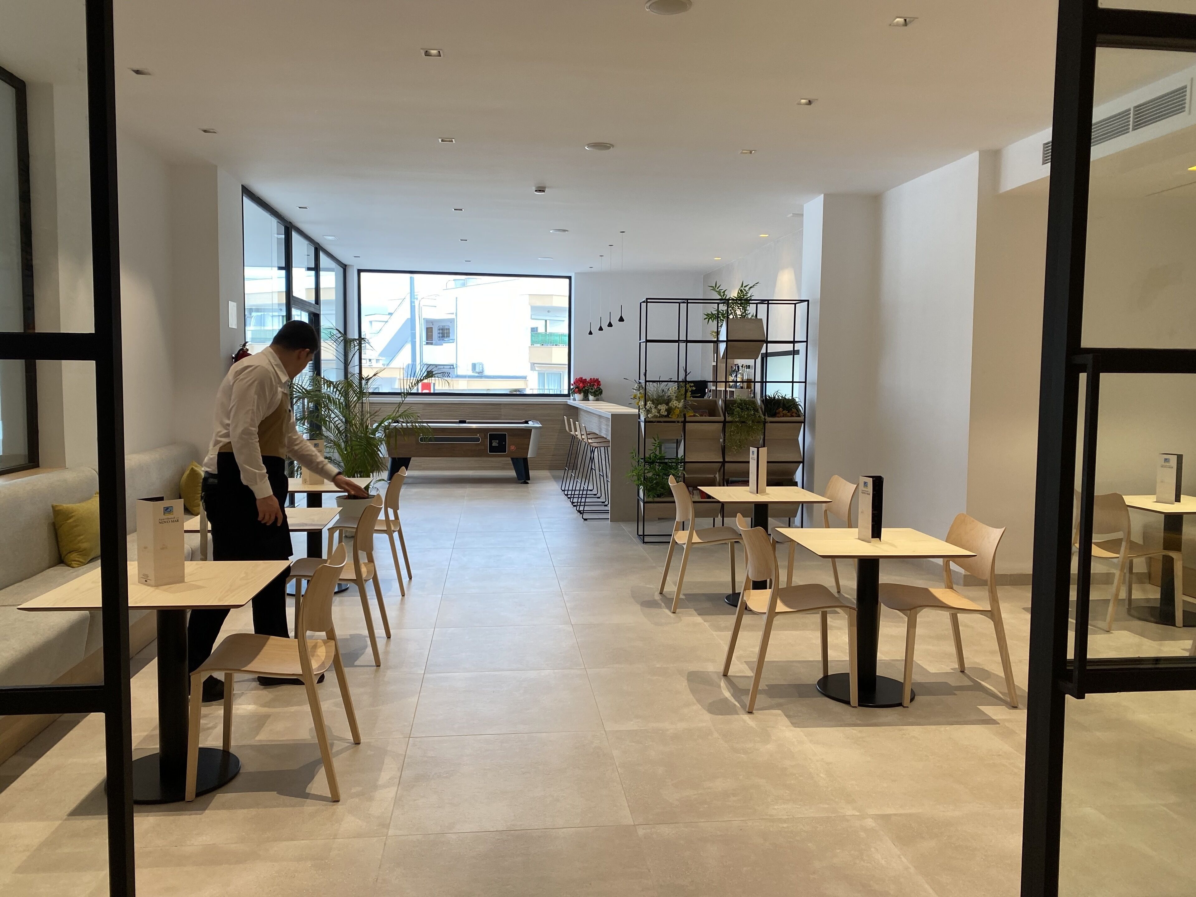 Un café moderno y luminoso con mesas y sillas de madera, un ventanal con vistas a la ciudad y una estantería con plantas al fondo.