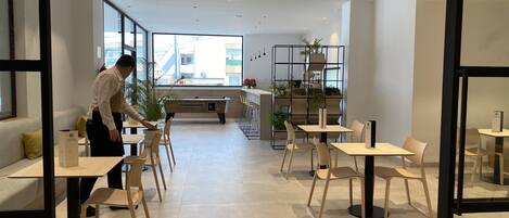 Un café moderno y luminoso con mesas y sillas de madera, un ventanal con vistas a la ciudad y una estantería con plantas al fondo.