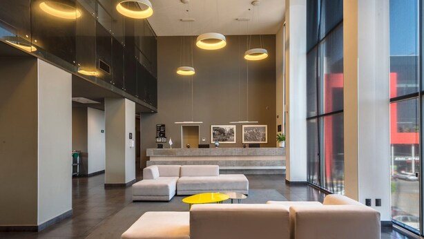 A spacious lobby with a modern design, featuring a large sectional sofa, a yellow coffee table, and a reception area with framed pictures and a counter. - City Express Plus by Marriott Guadalajara Palomar (Tlajomulco de Zúñiga)