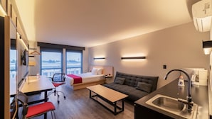 A compact hotel room with a bed, a sofa, a small table, a desk with a chair, a kitchenette, and a window with a view. - City Express Plus by Marriott Guadalajara Palomar (Tlajomulco de Zúñiga)