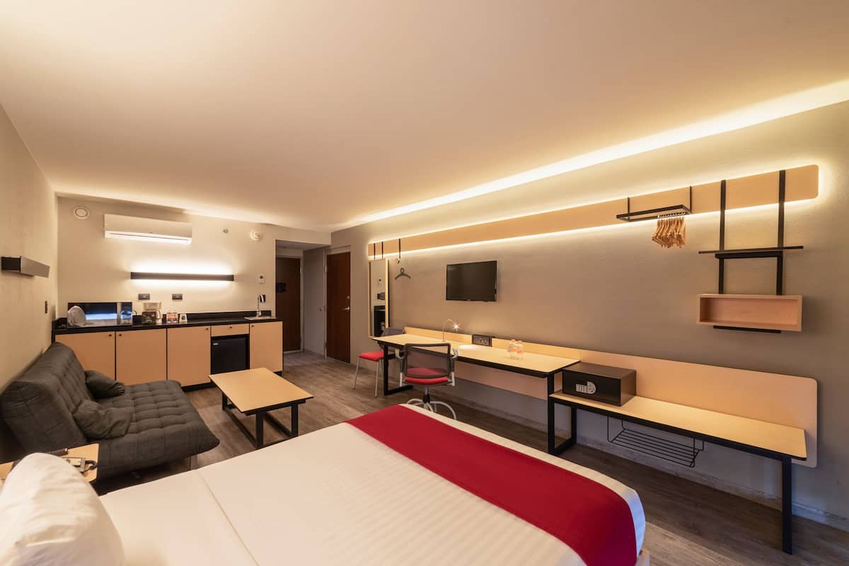 Ein modernes Hotelzimmer mit Bett, Schreibtisch, Sofa, einer kleinen Küchenzeile und einem an der Wand montierten Fernseher.