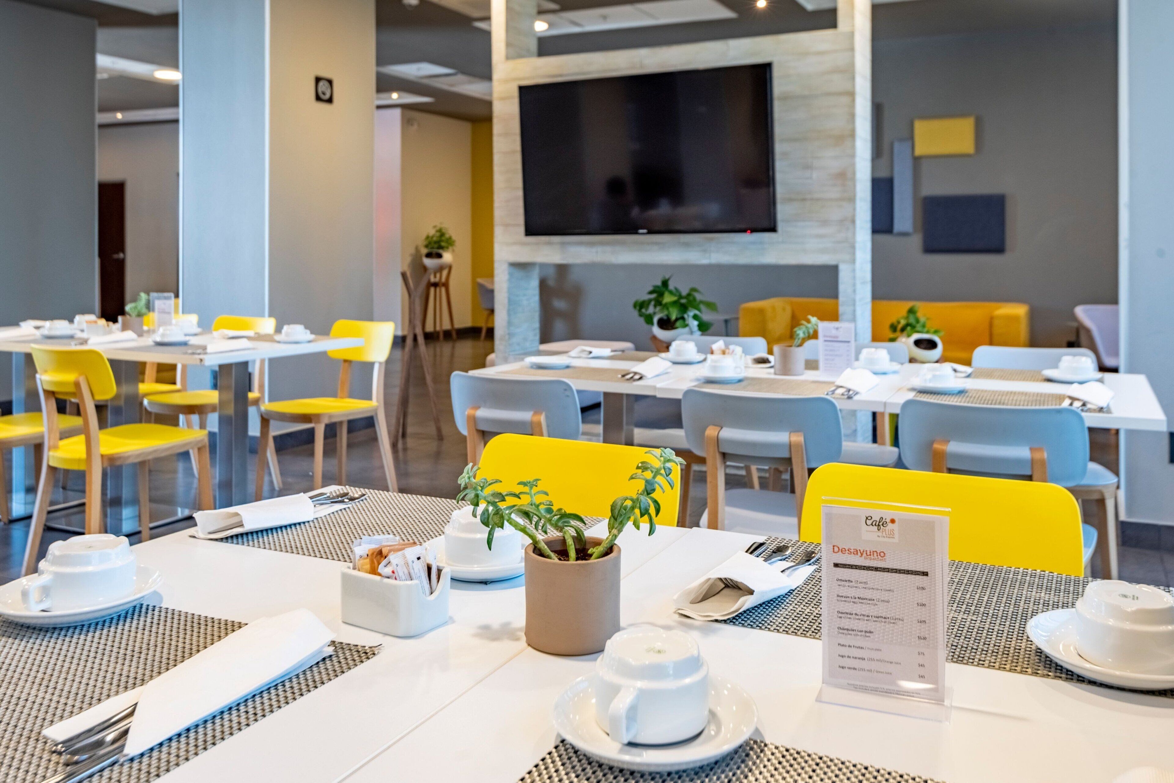 Un espace repas moderne, avec des tables préparées pour le petit-déjeuner, comprenant des chaises jaunes, de la vaisselle blanche et un présentoir à menus.