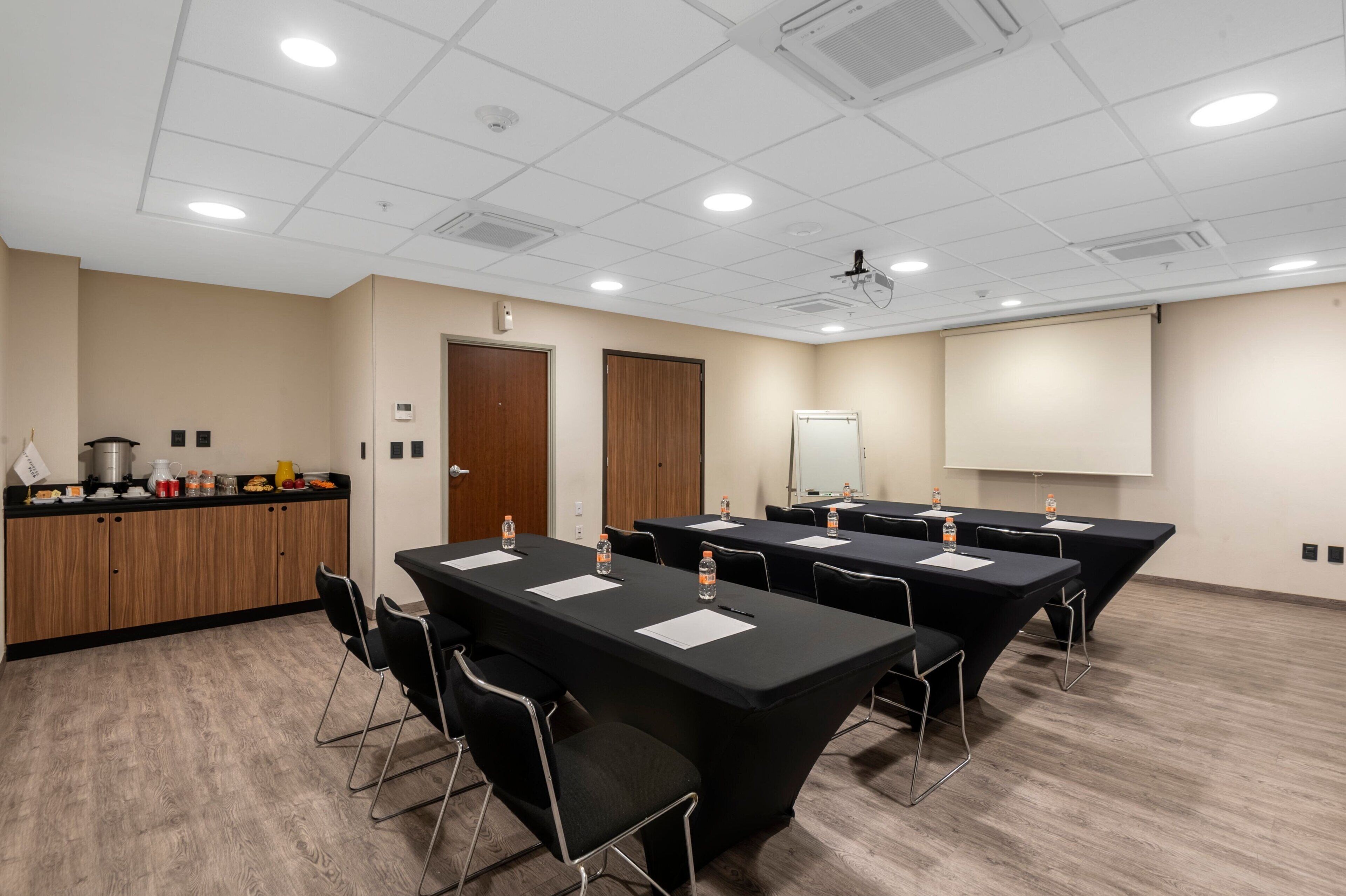 Une salle de conférence équipée de quatre tables, chacune avec des chaises, des bouteilles d’eau et des documents. On y trouve également une petite cuisine avec une cuisinière et des placards, ainsi qu’un écran de projection fixé au mur.