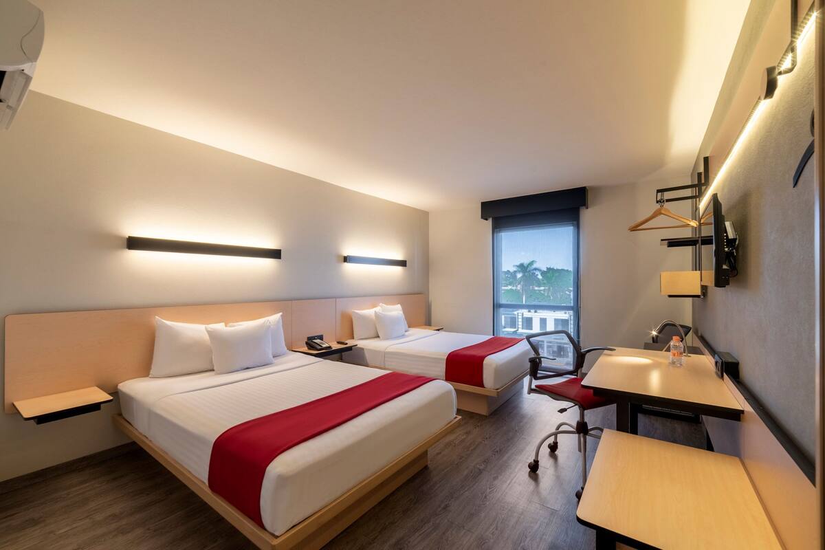 Ein Hotelzimmer mit zwei Betten, einem Schreibtisch, einem Stuhl, einem Fenster mit Blick und einem kleinen Tisch mit einer Flasche.