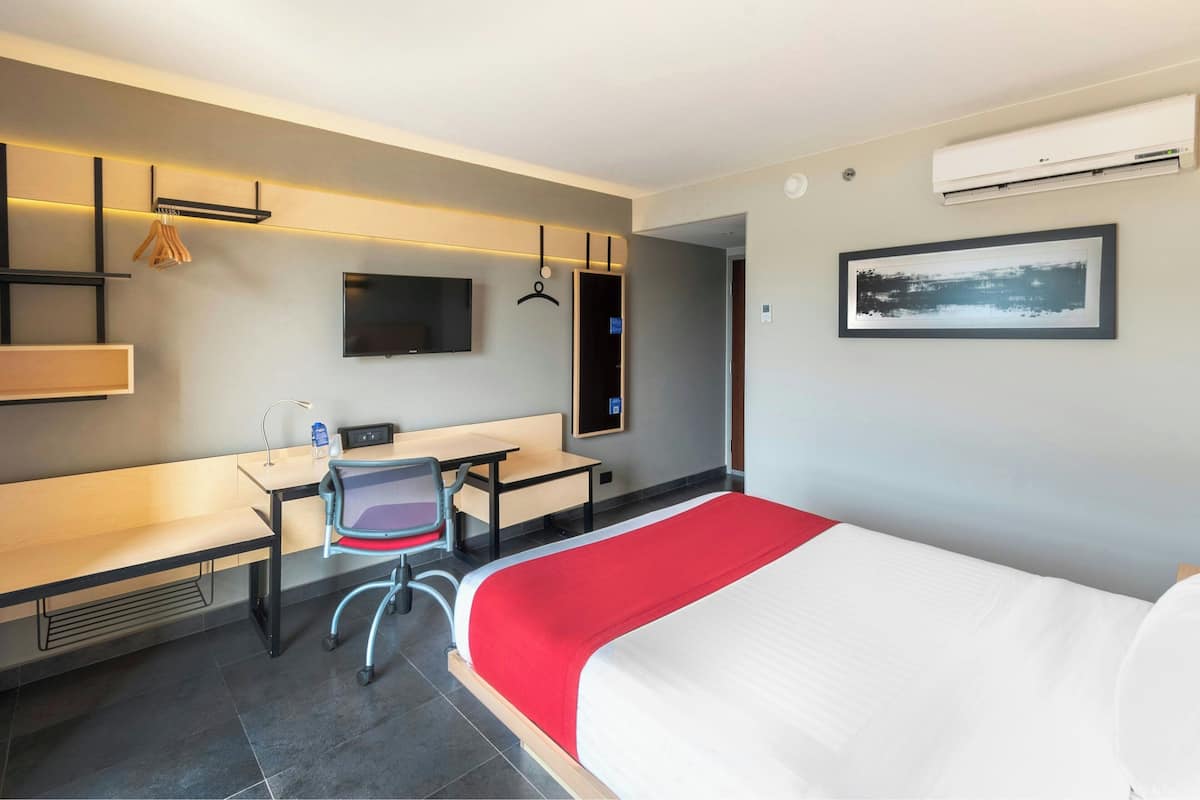 Ett modernt hotellrum med en säng, ett skrivbord, en stol, en platt-TV och en luftkonditioneringsanläggning.