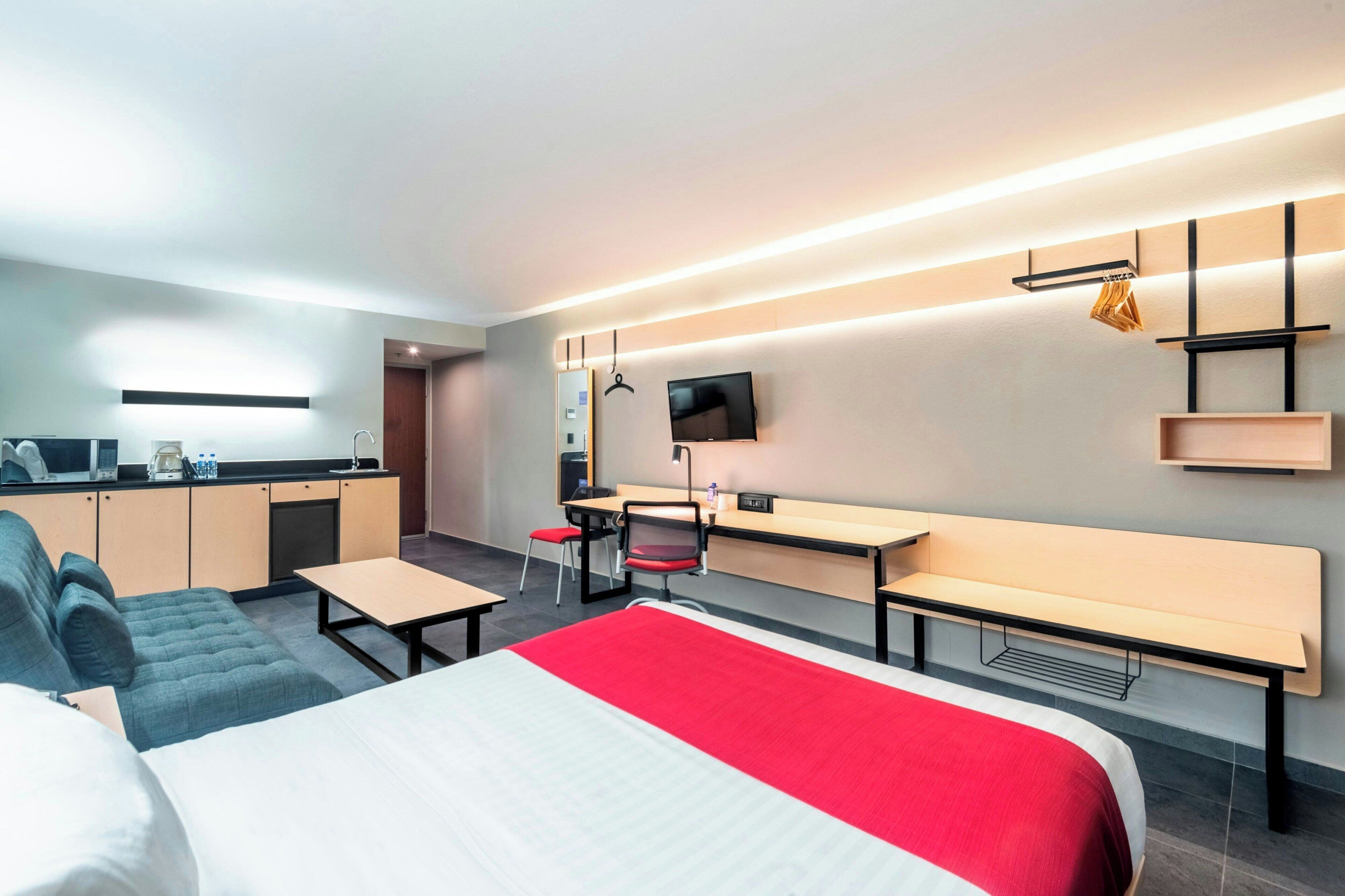A modern hotel room with a bed, a sofa, a desk, and a kitchenette.