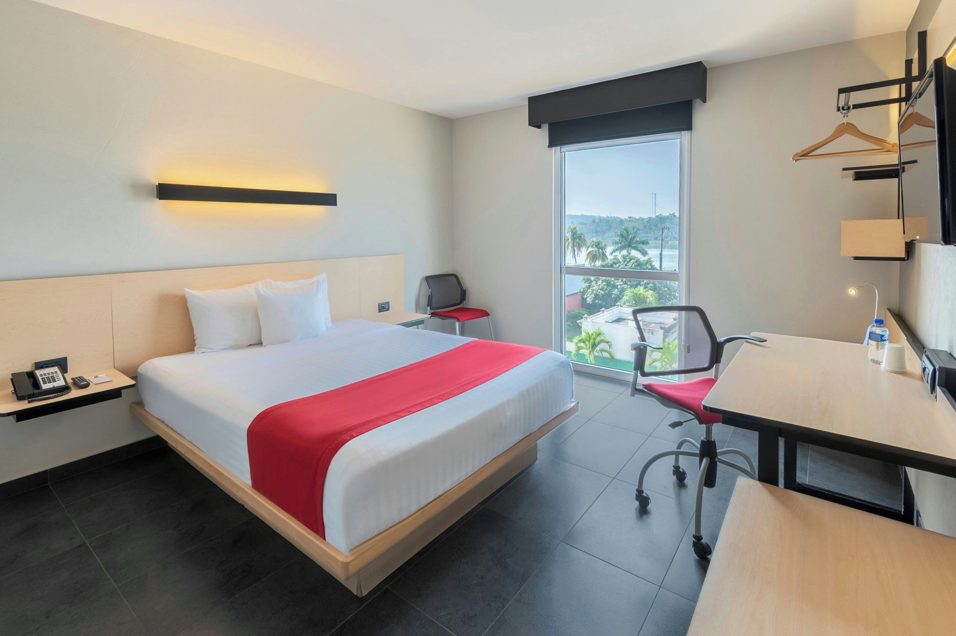 Habitación de hotel con una cama grande, un escritorio, una silla, una mesita, un teléfono, una botella de agua y una ventana con vistas.