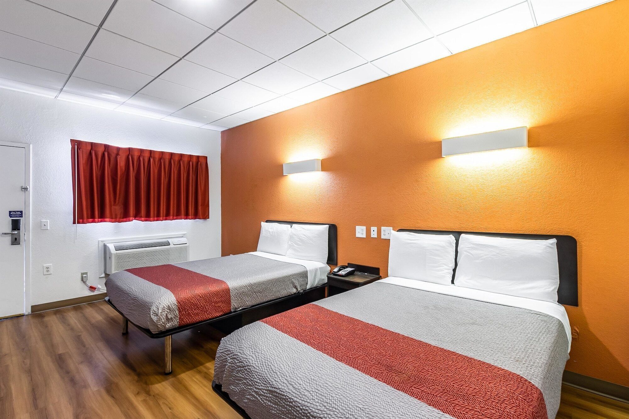 Une chambre d’hôtel avec deux lits, un rideau rouge, un appareil de climatisation et un luminaire fixé au mur.