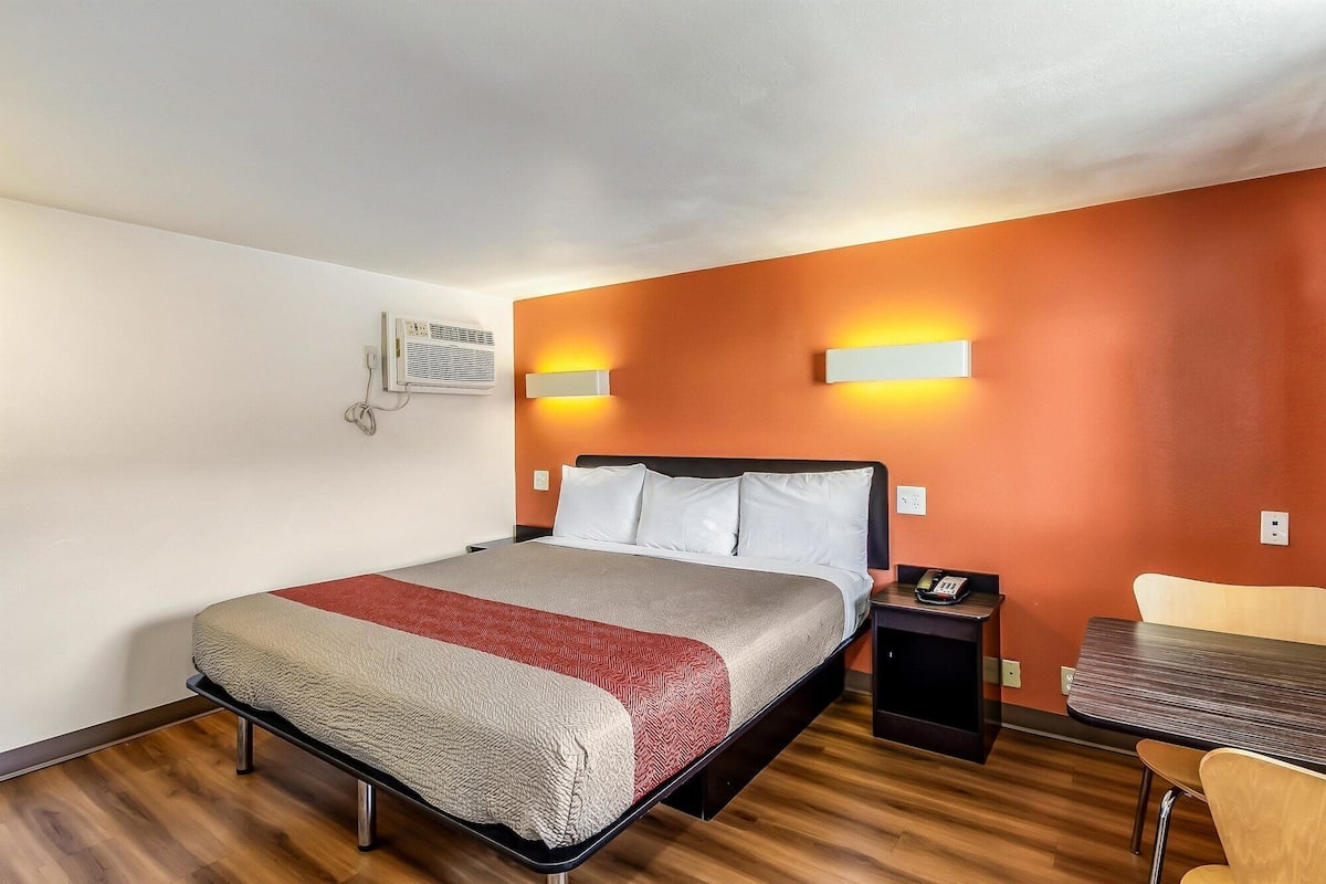 A hotel room with a bed, bedside table, chair, and a wall-mounted air conditioning unit.