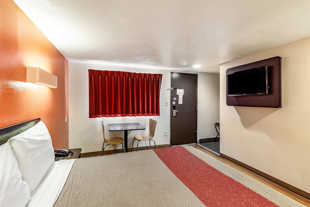 A hotel room with a bed, a desk, a chair, a TV mounted on the wall, and a door with a red curtain.