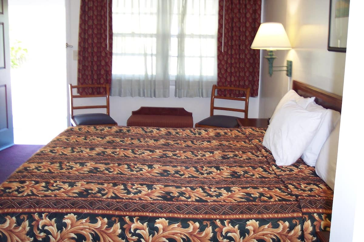 A hotel room with a large bed, two chairs, a window with curtains, and a bedside lamp.