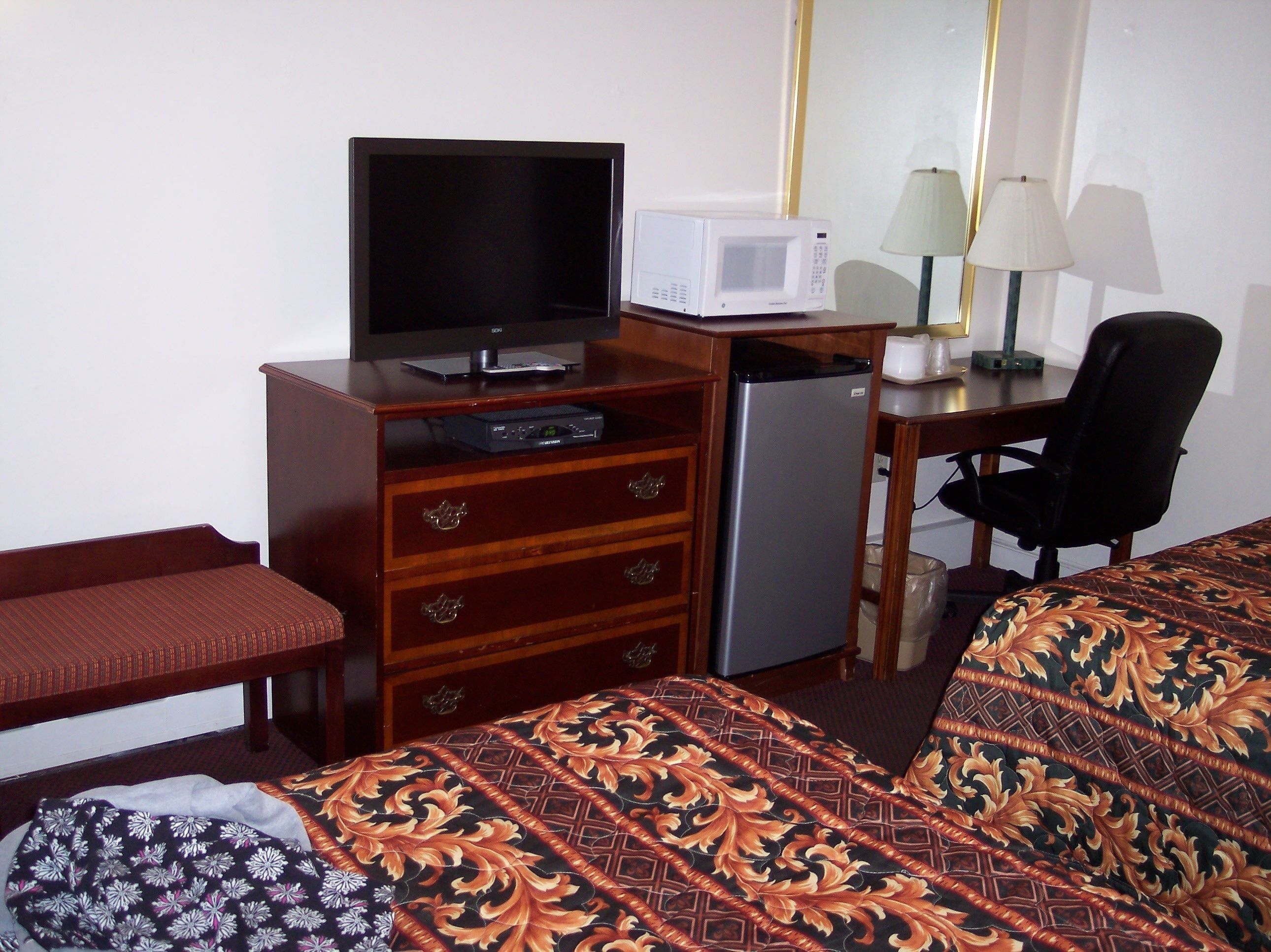 Habitación de hotel con cama, escritorio con microondas, televisor, refrigerador, una silla y una lámpara.