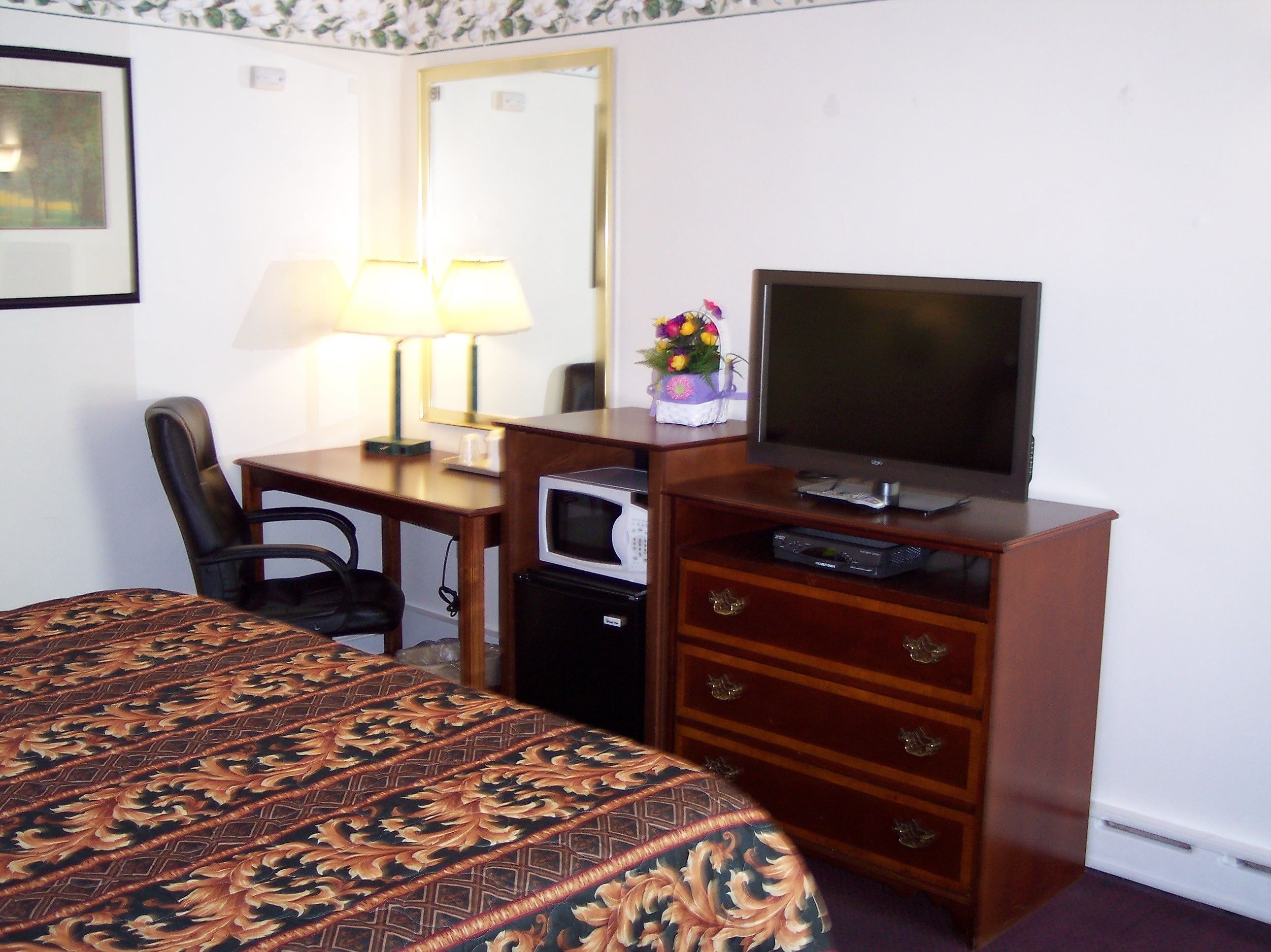 A hotel room with a bed, a desk with a lamp, a television, a microwave, and a dresser with drawers.