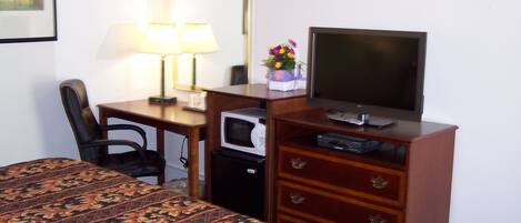 A hotel room with a bed, a desk with a lamp, a television, a microwave, and a dresser with drawers.