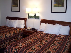 Habitación de hotel con dos camas, cada una con una colcha estampada y almohadas blancas. Un mesita de noche entre las camas tiene un teléfono y un pequeño objeto negro. Dos lámparas de pared brindan iluminación.