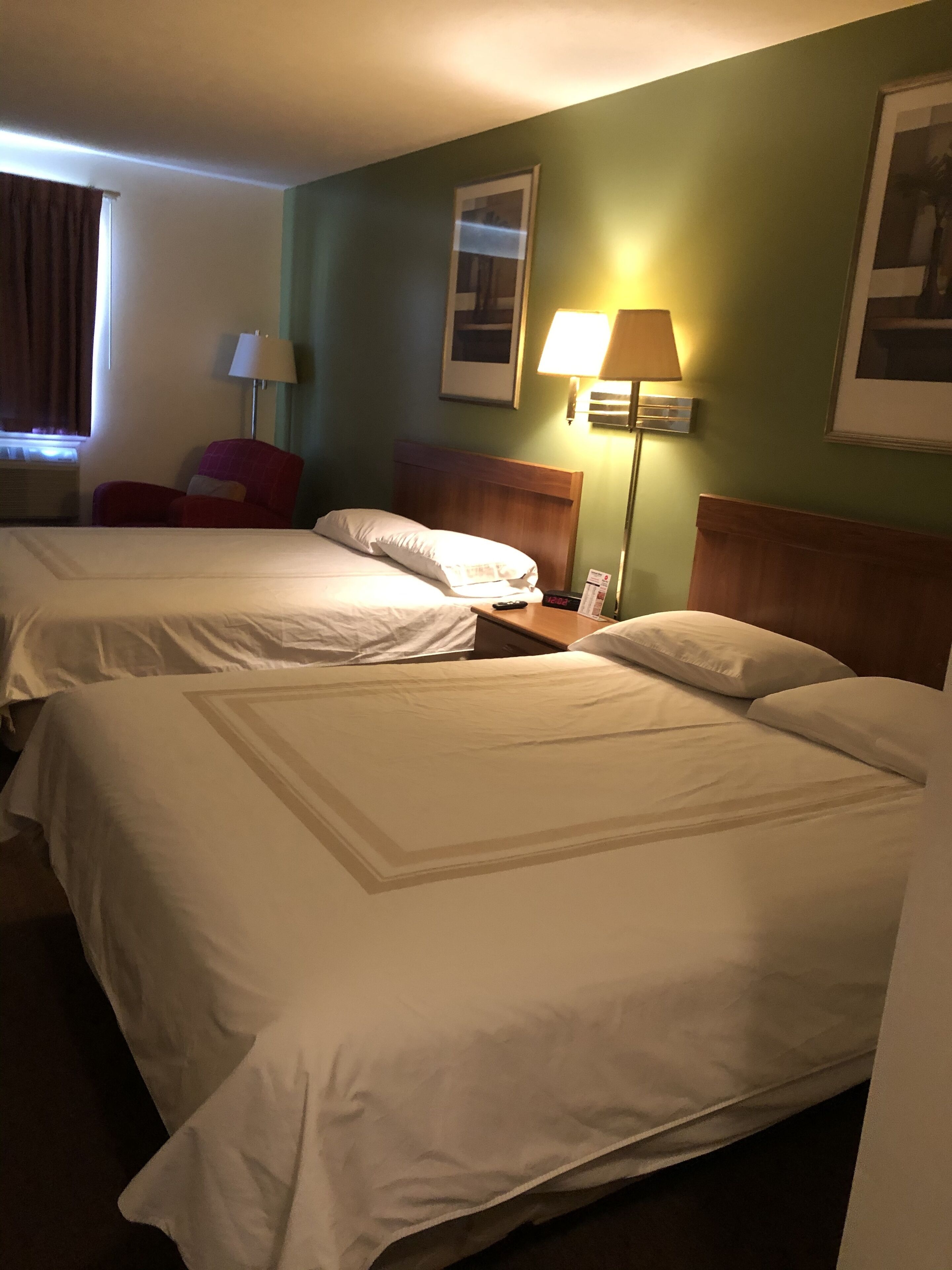 A hotel room with two beds, a chair, a nightstand, a lamp, and framed pictures on the wall.