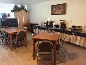 Uma sala de jantar com mesa e cadeiras de madeira, bancada de cozinha com eletrodomésticos e um quadro na parede.