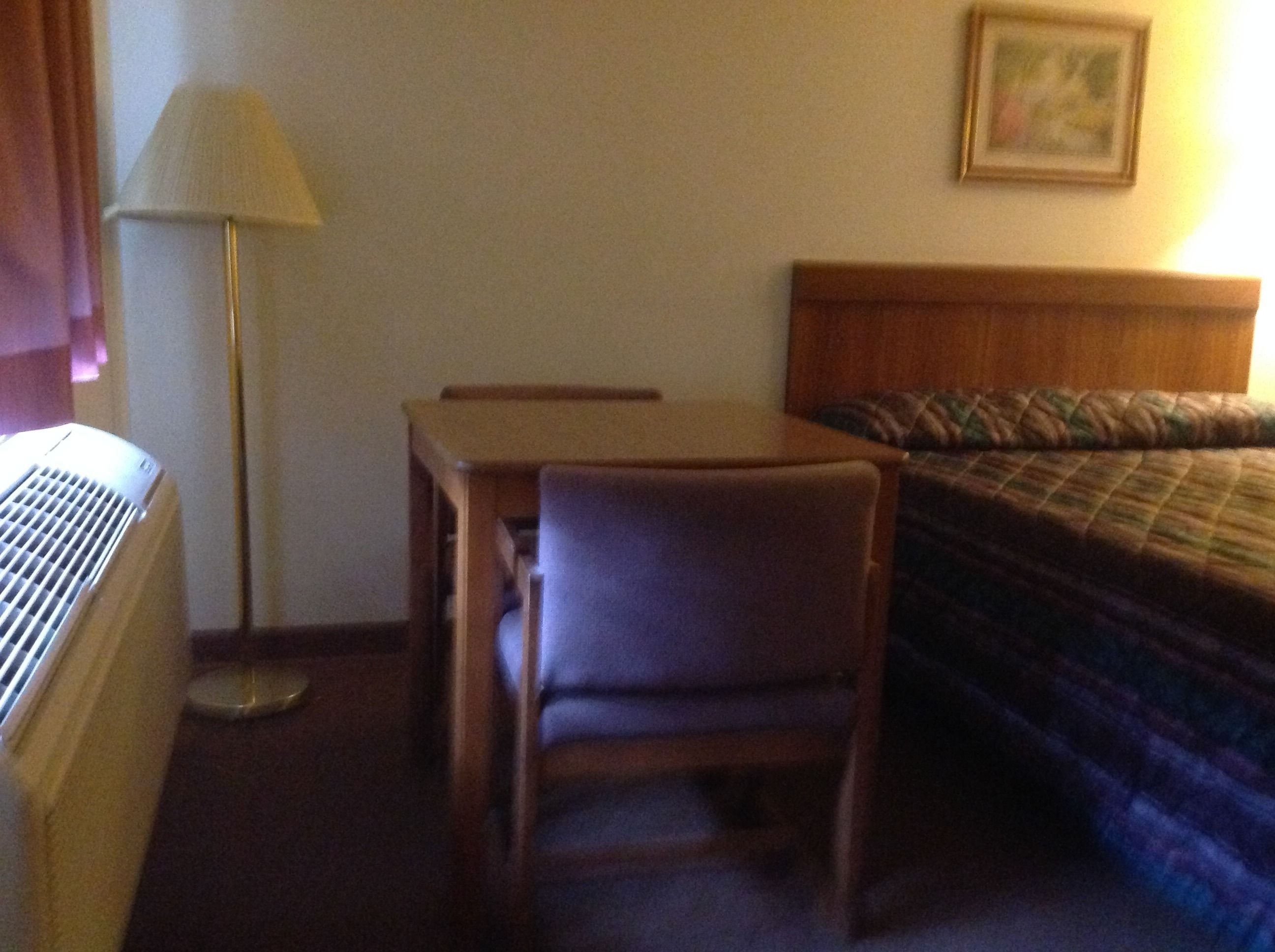 Quarto de hotel pequeno com uma cama de solteiro, uma escrivaninha, uma cadeira, um abajur e um radiador.
