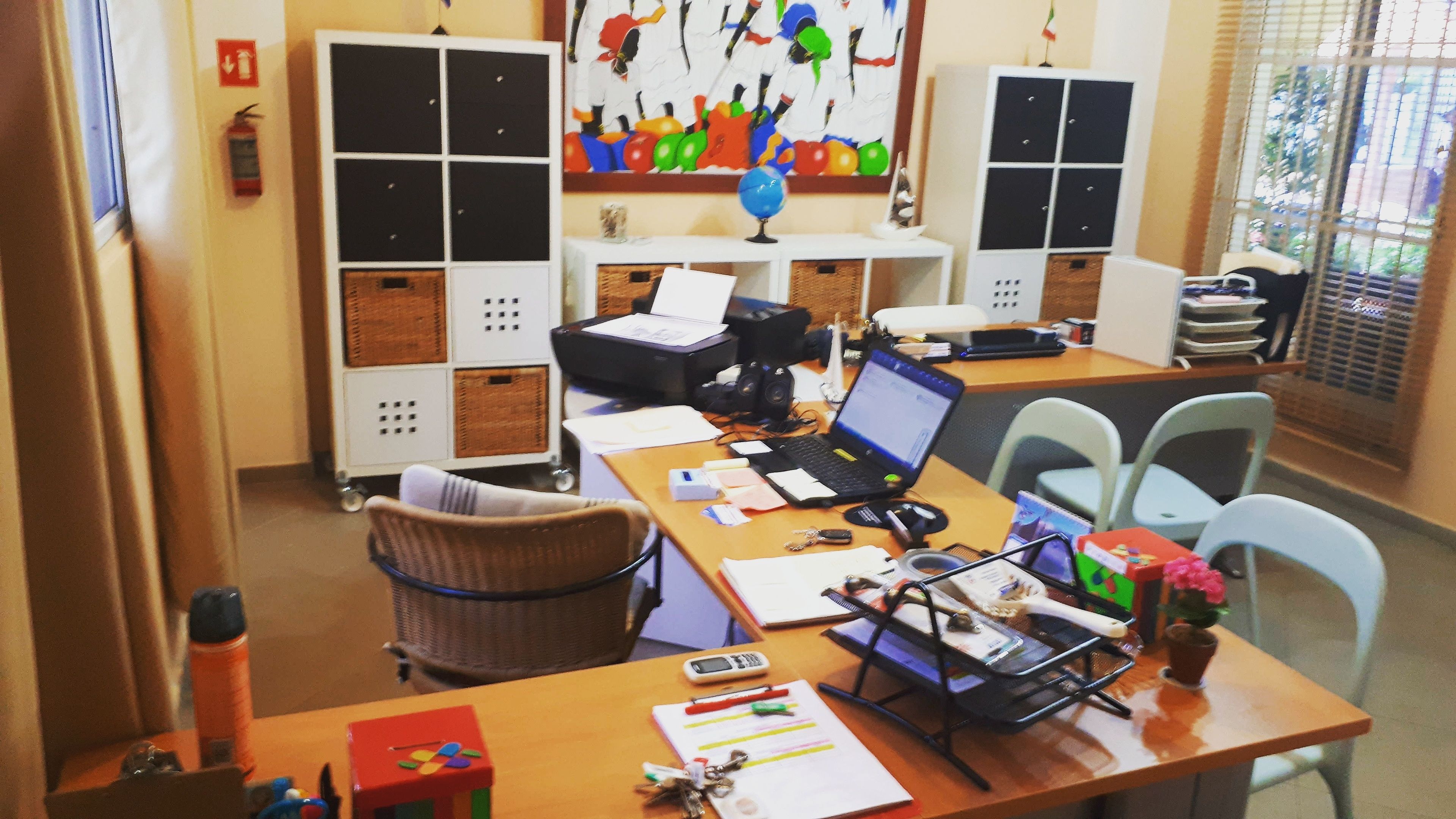Uma sala de aula com uma mesa, cadeiras, um laptop, uma impressora e armários.