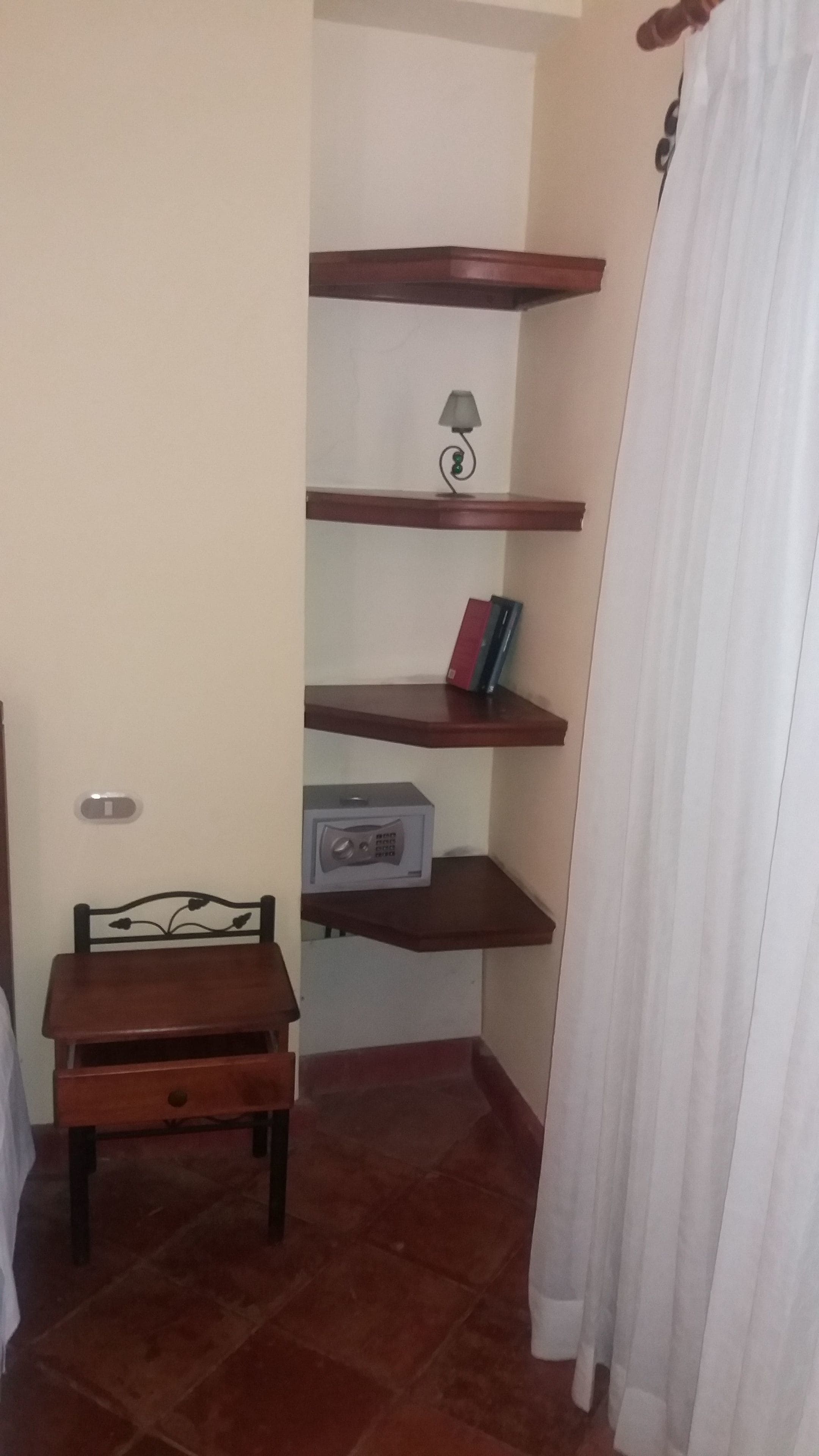 A corner room with a small wooden chair, a radio, and a shelf with books and a lamp.