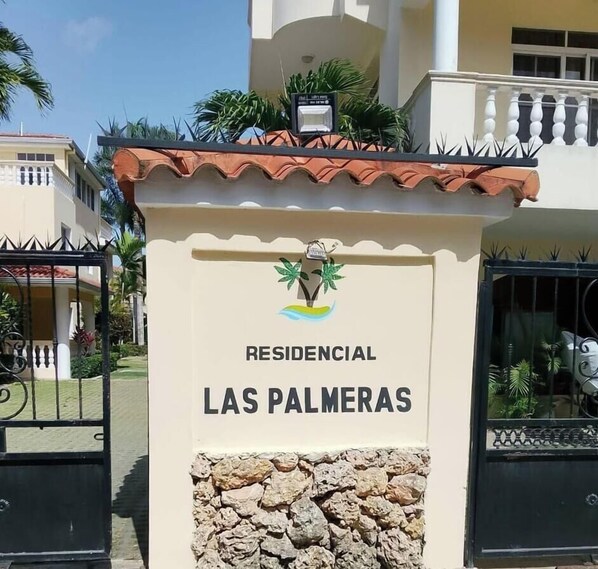Une entrée fermée par un portail, sur laquelle on peut lire l’inscription « RESIDENCIAL LAS PALMERAS », et un petit logo au-dessus.