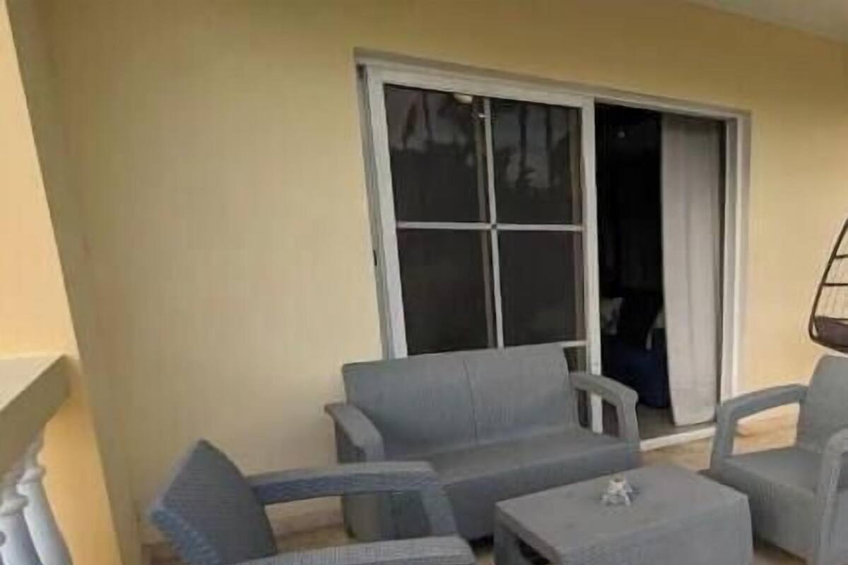 Ein Balkon mit grauer Gartenmöbeln, ein Fenster mit Jalousien und Blick ins Innere.