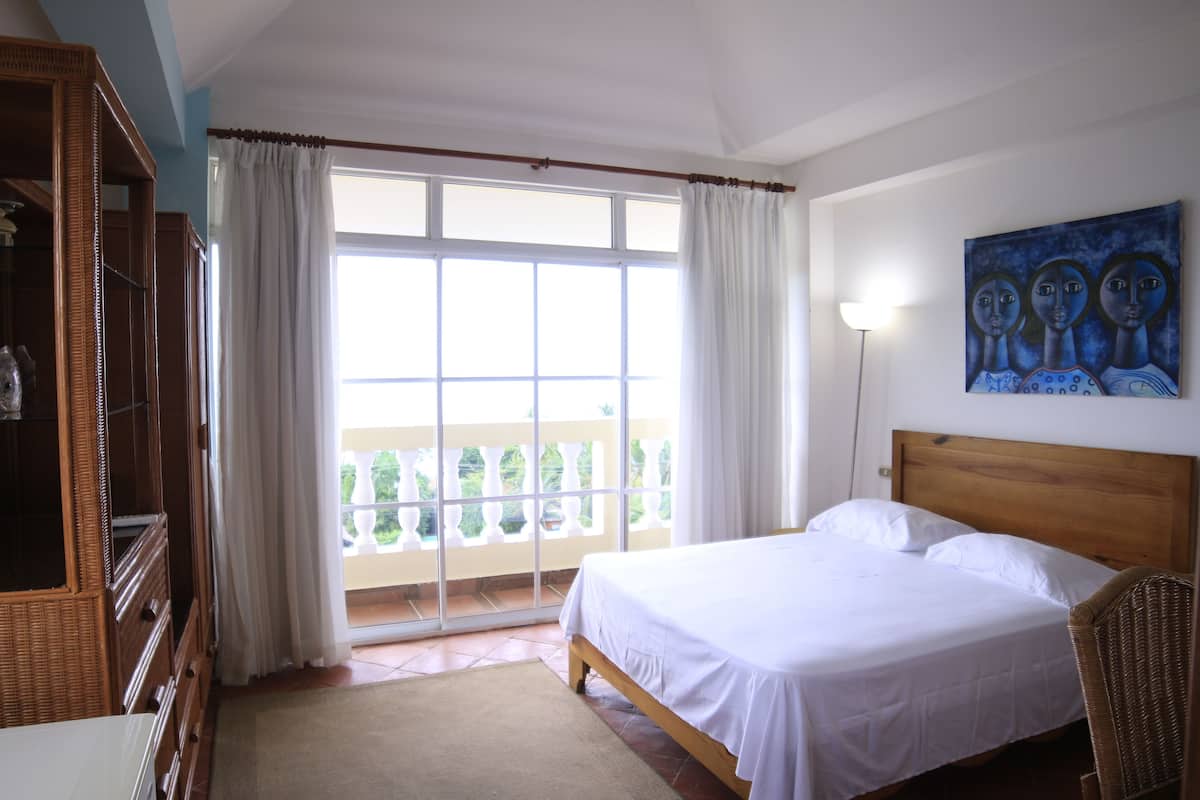 Ein Schlafzimmer mit großem Fenster, einem Bett, einem Sessel und einem Gemälde an der Wand.