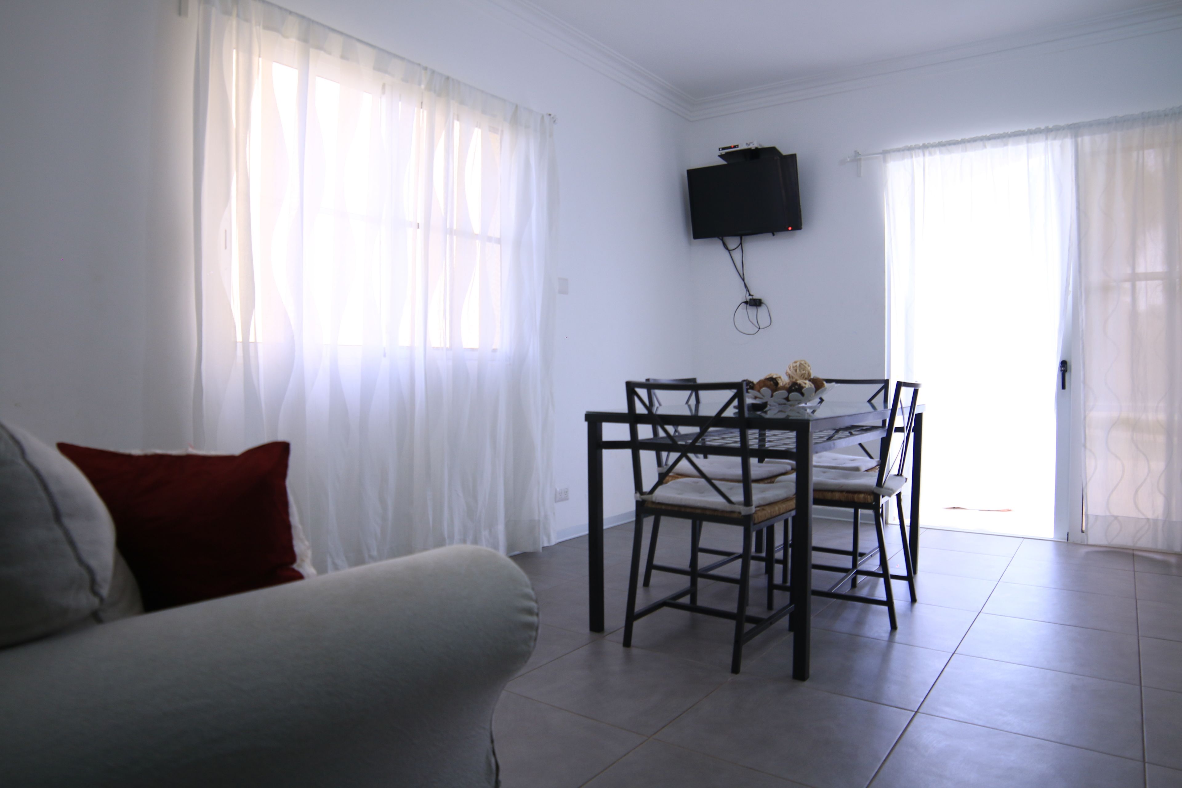 A living room with a sofa, a dining table with chairs, a TV mounted on the wall, and a door leading outside.