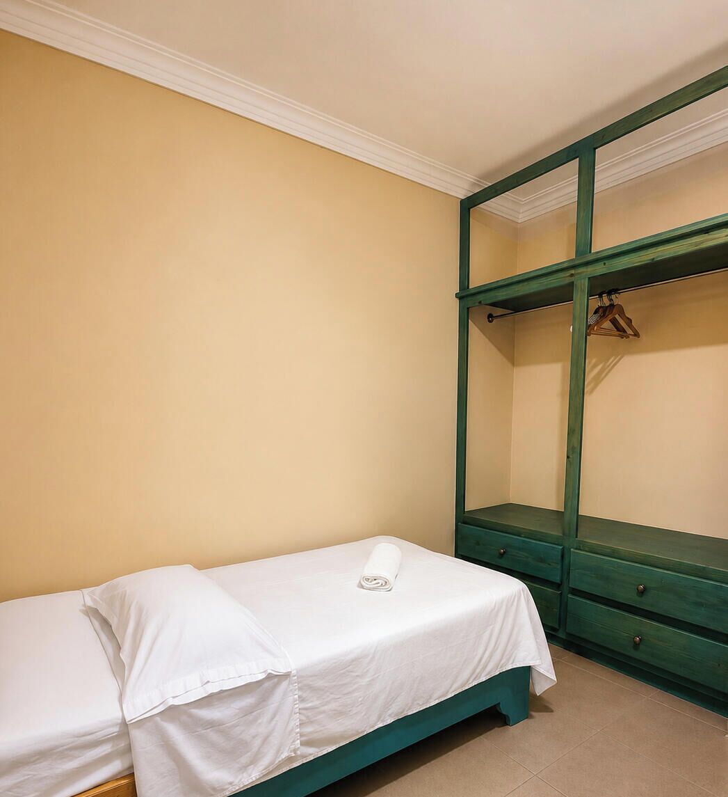 Una cama individual con ropa de cama blanca en una habitación con un armario verde y un perchero blanco.