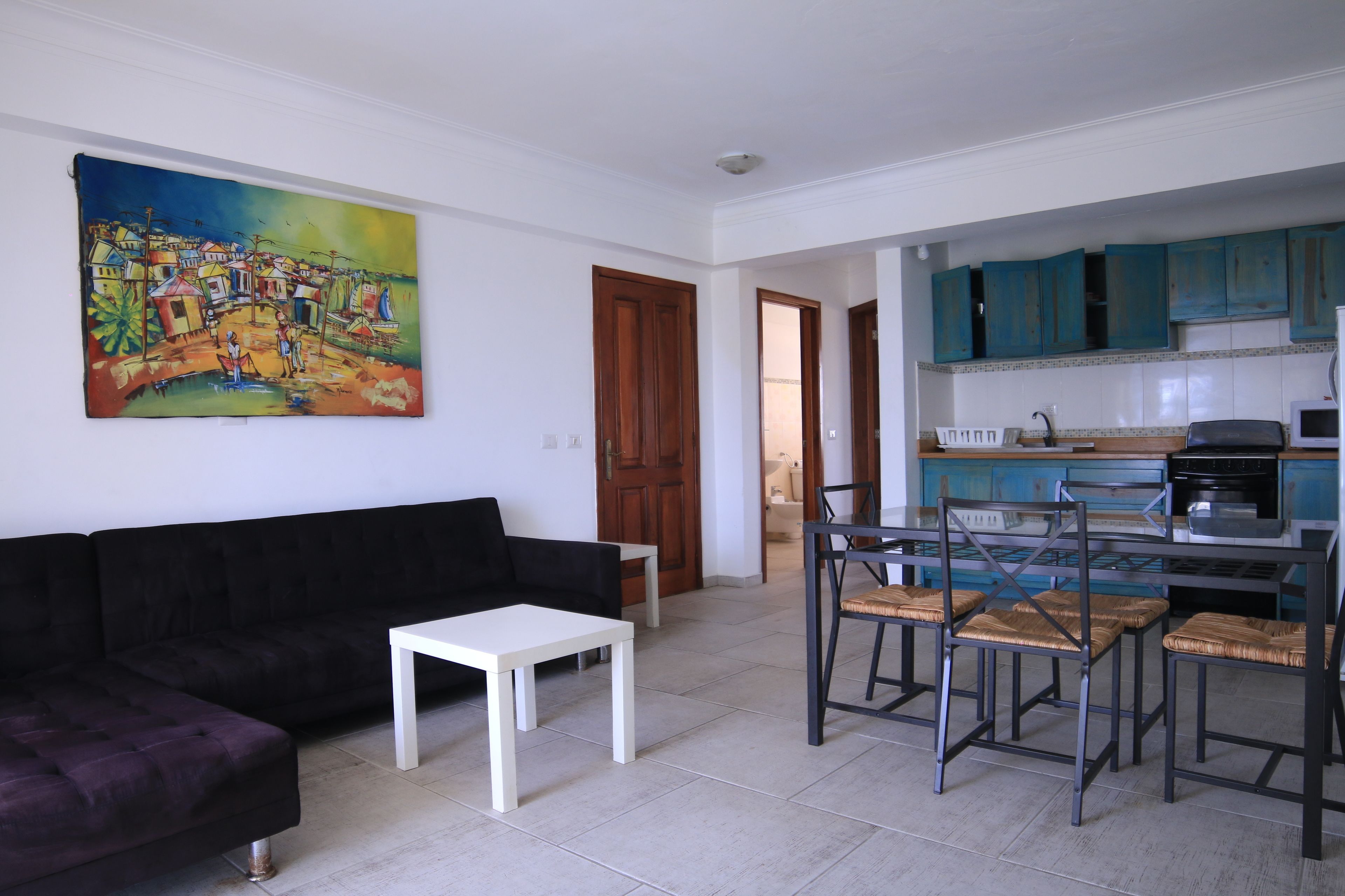 Una sala de estar con un sofá negro, una mesa de centro blanca y una zona de comedor con una mesa de metal y sillas de mimbre. Hay un cuadro colorido en la pared y una cocina con armarios azules.