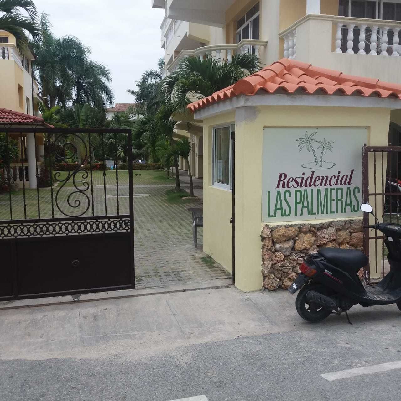 A gated entrance to a residential complex named 'Las Palmeras' with a paved pathway leading to the entrance.