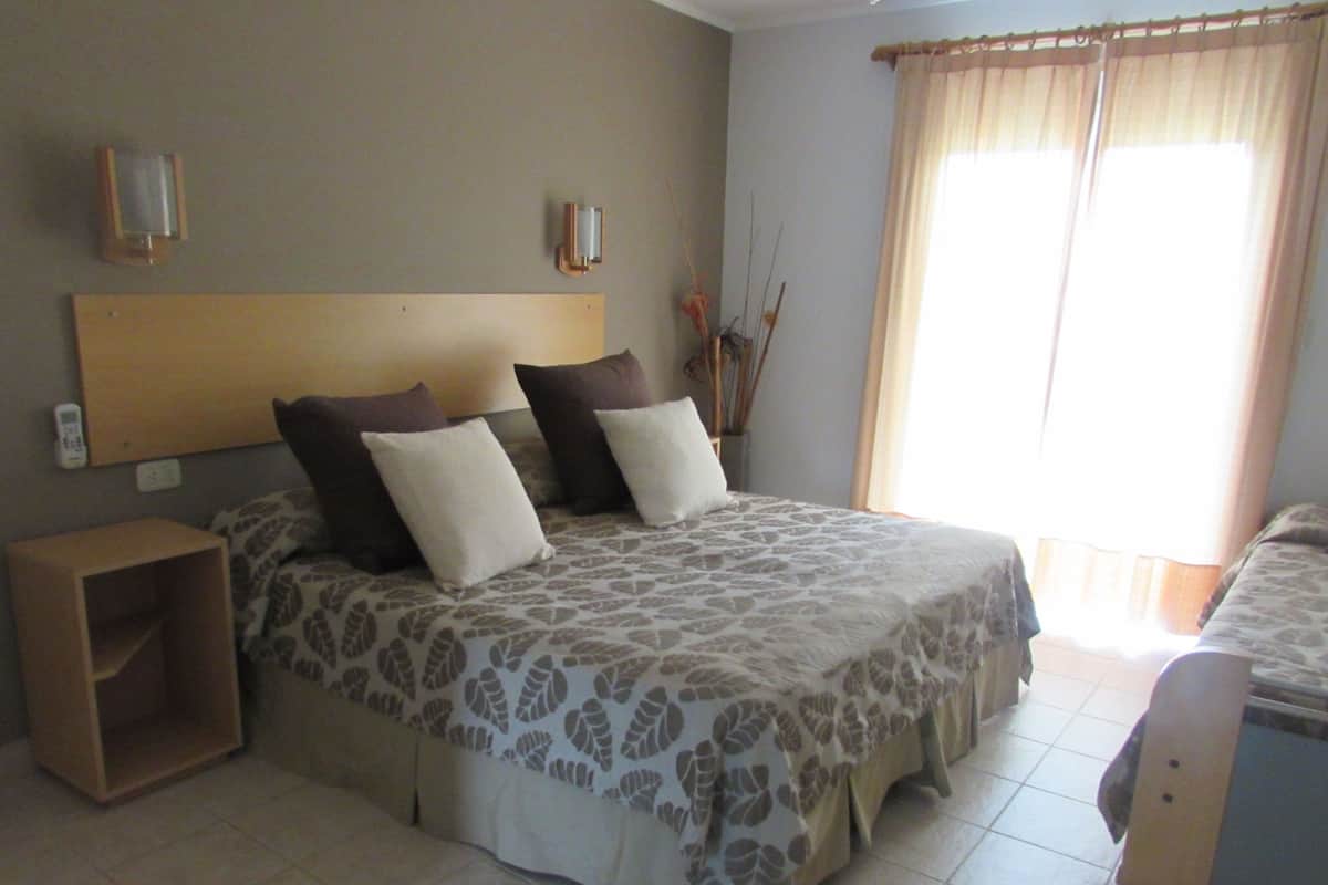 A bedroom with a bed, bedside tables, a nightstand, a wall-mounted light, and a window with curtains.