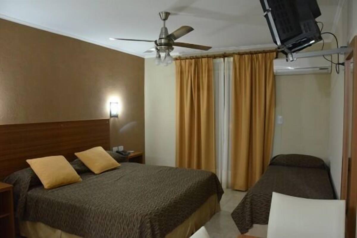 A hotel room with a bed, a smaller bed, a ceiling fan, a TV mounted on the wall, and a window with curtains.