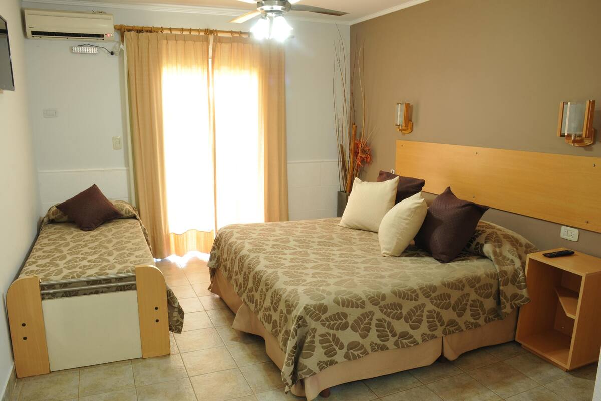 A bedroom with two beds, a ceiling fan, an air conditioning unit, a bedside table, and a window with curtains.