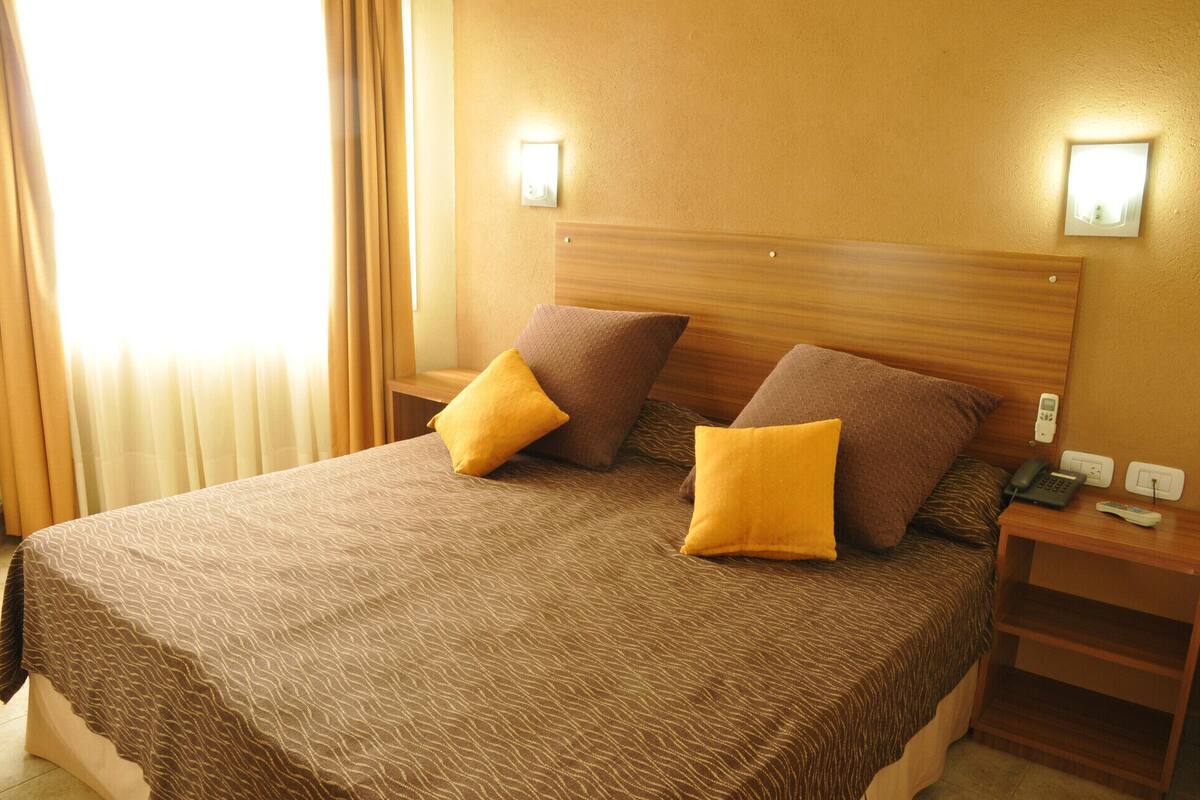 A hotel room with a bed, two pillows, a nightstand, and a wall-mounted light.