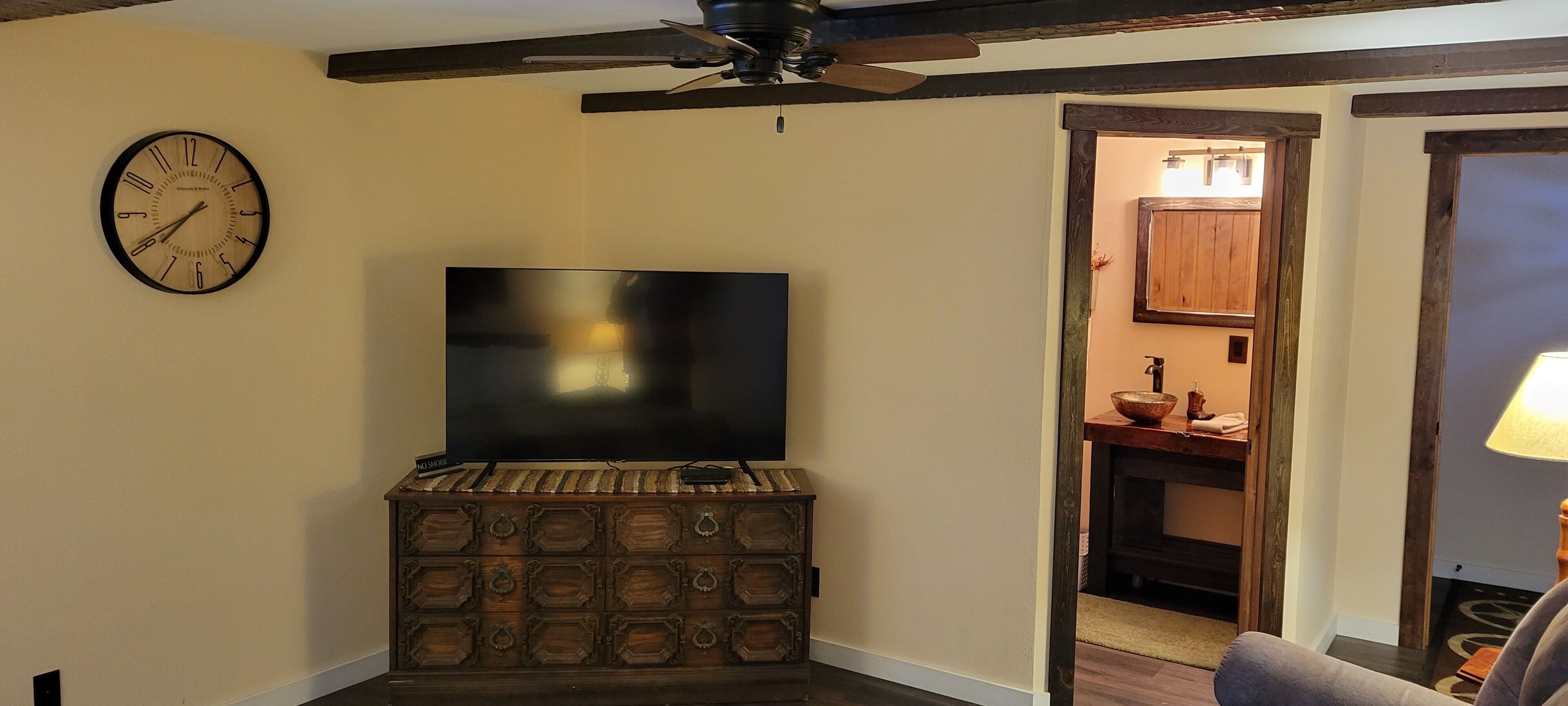Habitació amb televisió de pantalla plana penjada sobre un arca de fusta, ventilador de sostre i porta de fusta que dona a un bany.