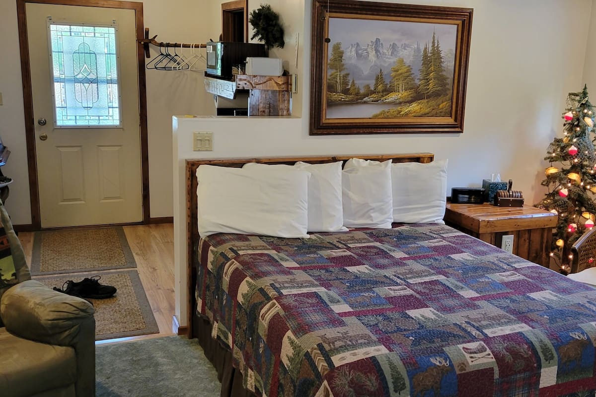 A bedroom with a bed, a chair, a Christmas tree, a painting, and a window with an air conditioner.