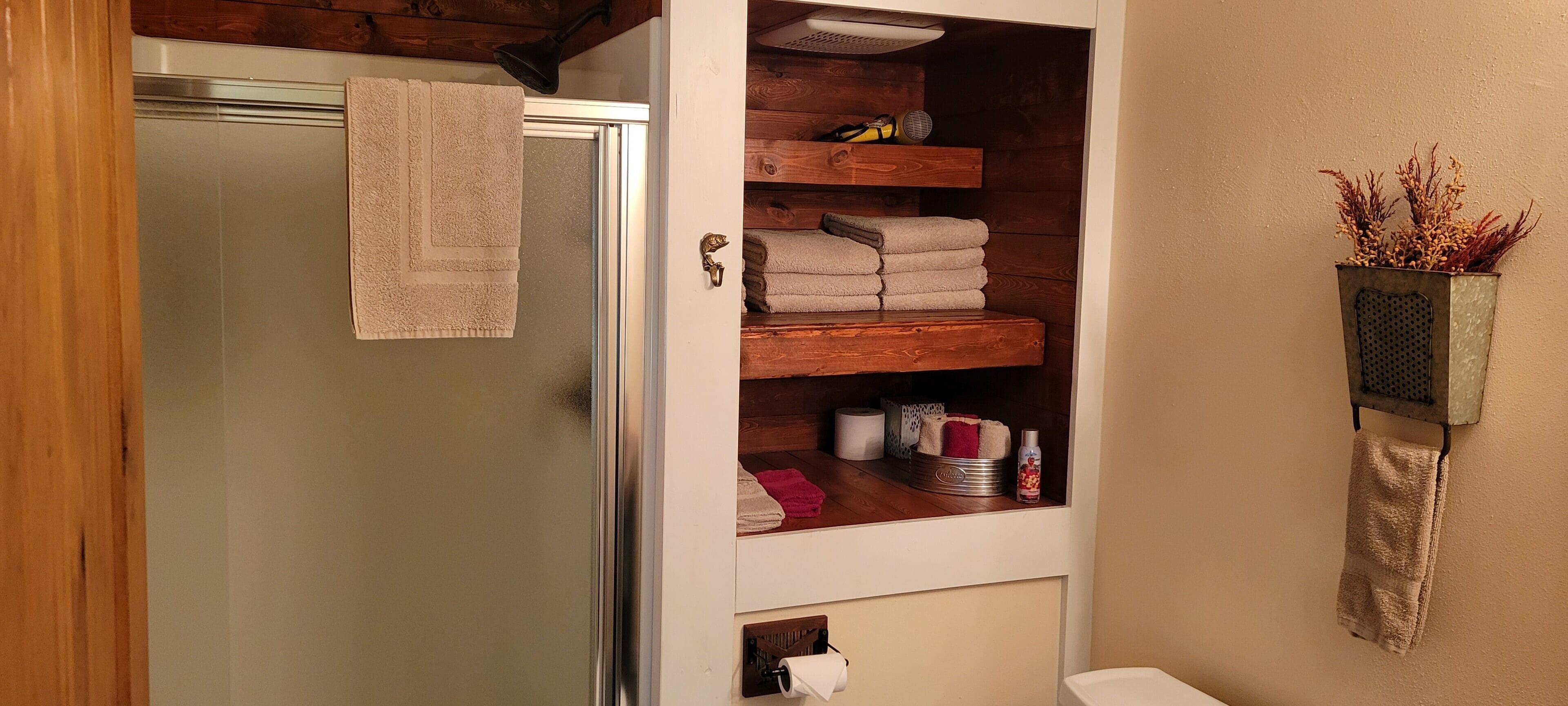 A bathroom with a wooden shelf holding towels and toiletries, a towel hanging on the door, and a small plant on the wall.