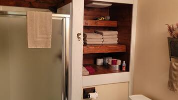 A bathroom with a wooden shelf holding towels and toiletries, a towel hanging on the door, and a small plant on the wall.