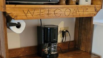 Una kitchenette compacta con microondas, cafetera y refrigerador pequeño. Un basurero se encuentra debajo de la encimera, y un cartel con la palabra 'WELCOME' está exhibido en el estante de madera superior.