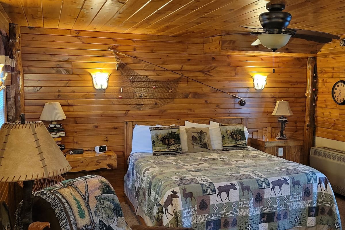 A cozy log cabin bedroom with a bed, two wall-mounted lamps, a ceiling fan, a clock, and a window with blinds.
