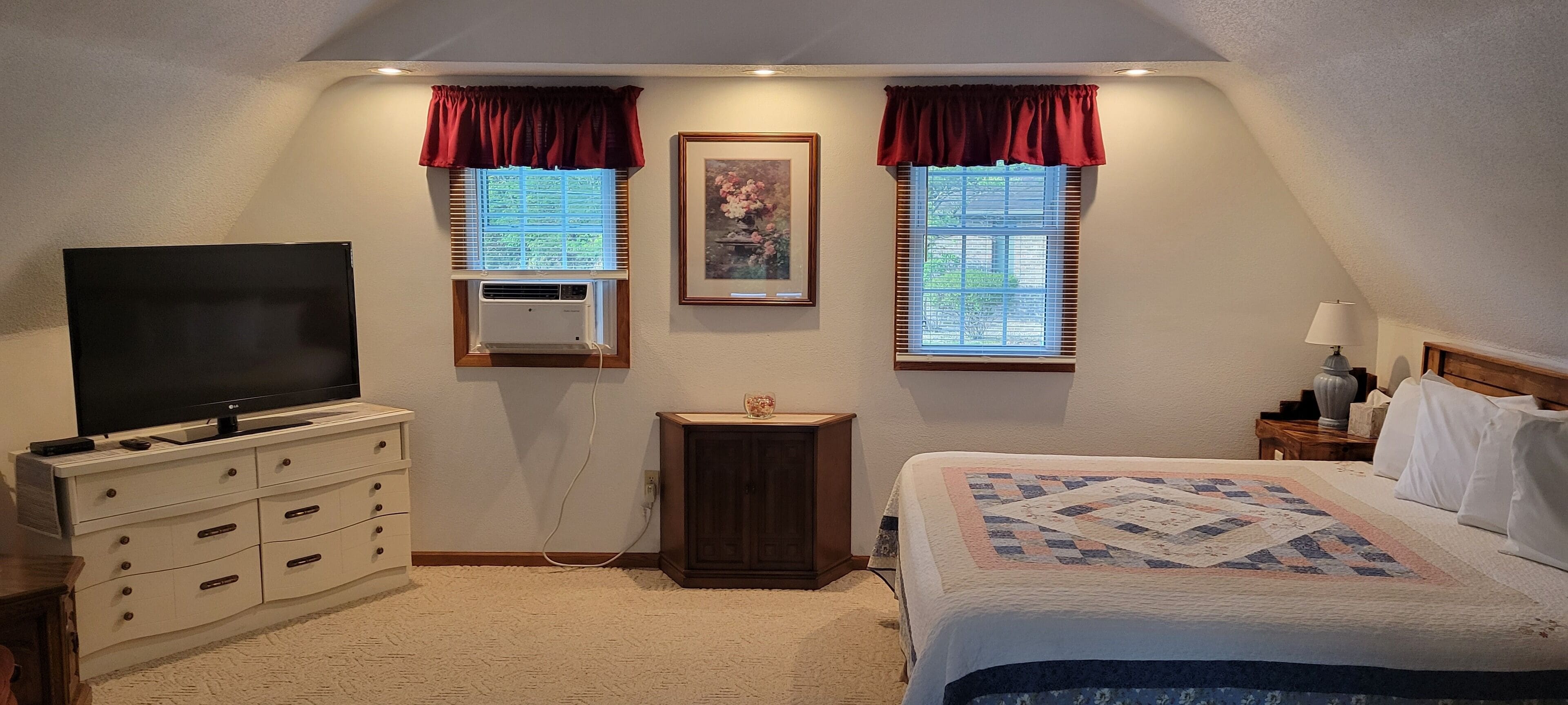 A bedroom with a bed, a dresser, a TV, two windows with curtains, a small table, and a nightstand.