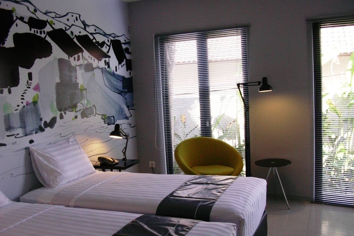 A modern hotel room with two beds, a yellow chair, a floor lamp, and a large window with blinds.